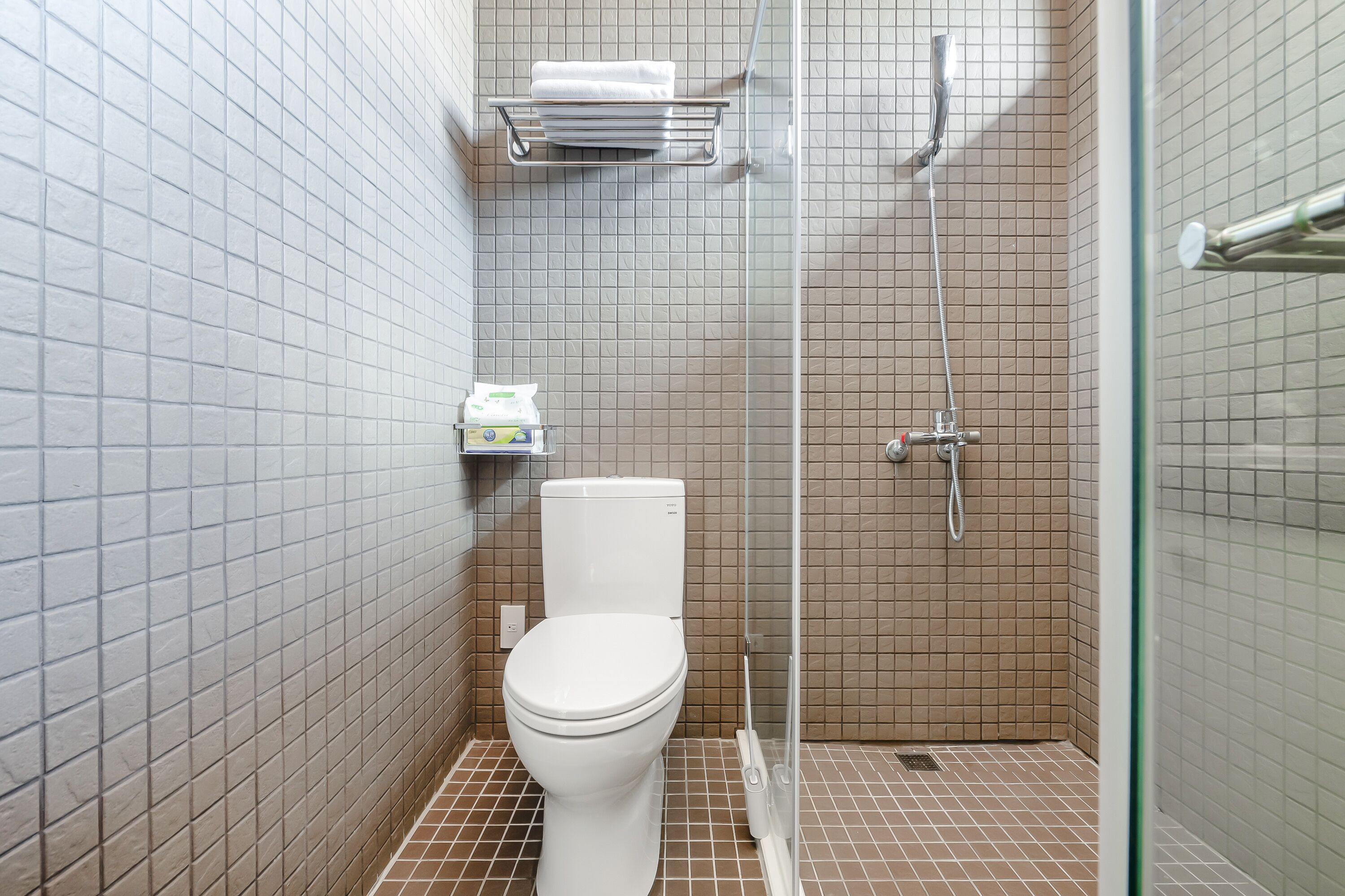 A small, tiled bathroom with a toilet, a shower, and a shelf with toiletries.