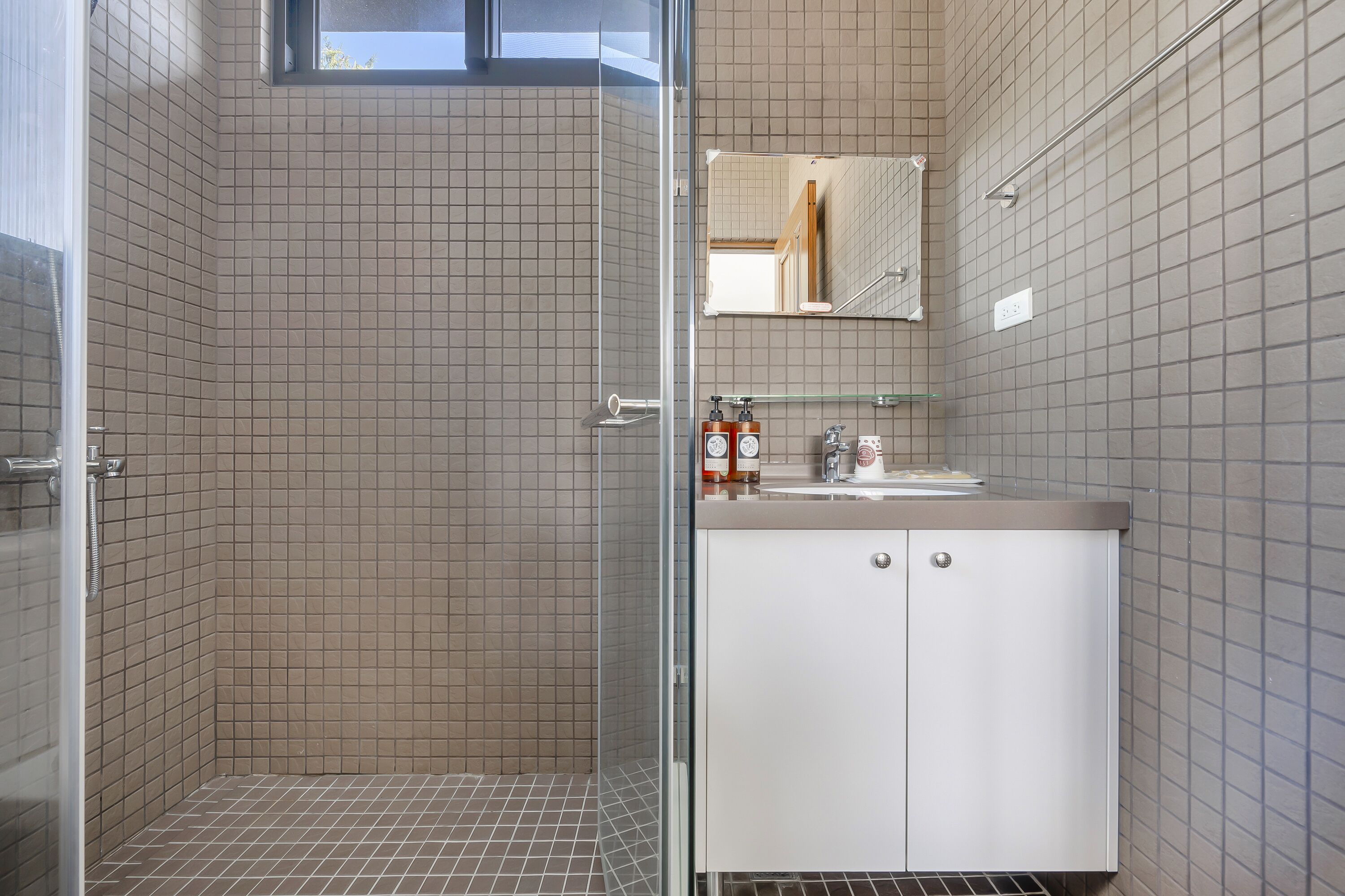 A modern bathroom with a glass shower enclosure, a white sink with a mirror above, and a tiled floor.
