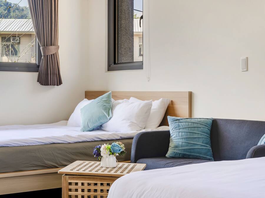 Une chambre à coucher avec un lit, un canapé, une petite table sur laquelle est posé un vase de fleurs, et une fenêtre avec des rideaux.