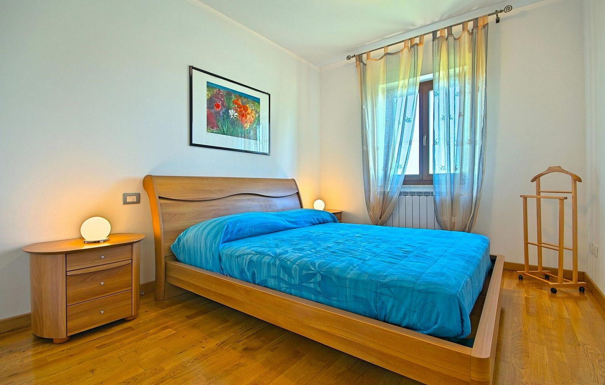 A bedroom with a wooden bed, a bedside table with a lamp, a framed picture on the wall, and a window with sheer curtains.