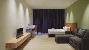 A modern hotel room with a flat-screen TV on a wooden console, a bed with a dark headboard, a grey sofa, and a large window with dark curtains. - Roxy Beach Apartment (Lundu)