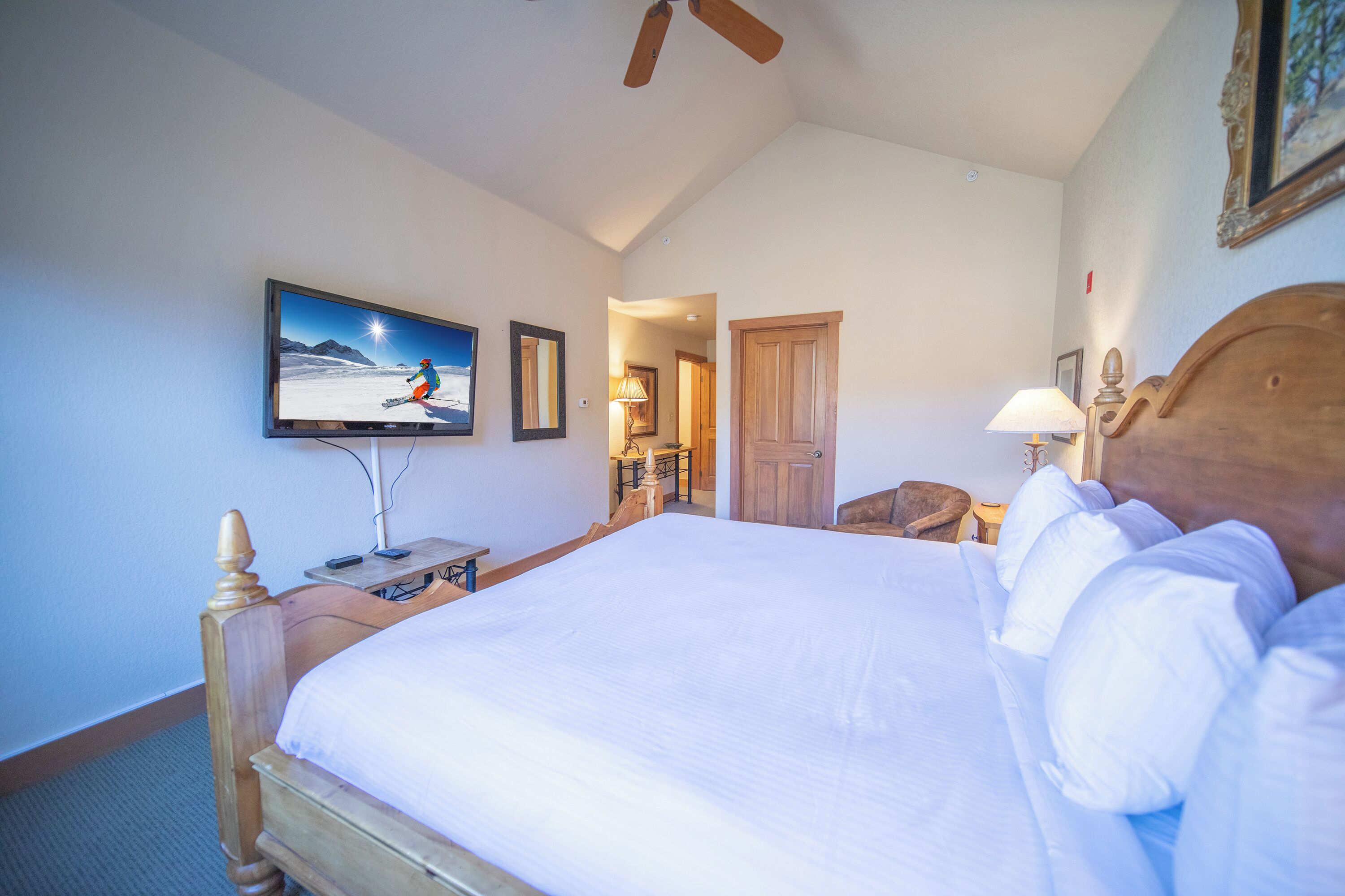 Ein Schlafzimmer mit einem großen Bett, einem an der Wand montierten Fernseher, einem hölzernen Kopfteil und einem Deckenventilator.