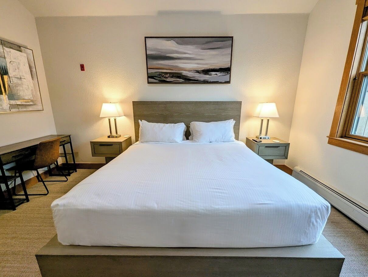 A neatly made bed in a hotel room with a wooden headboard, two bedside tables with lamps, a desk, and a chair.