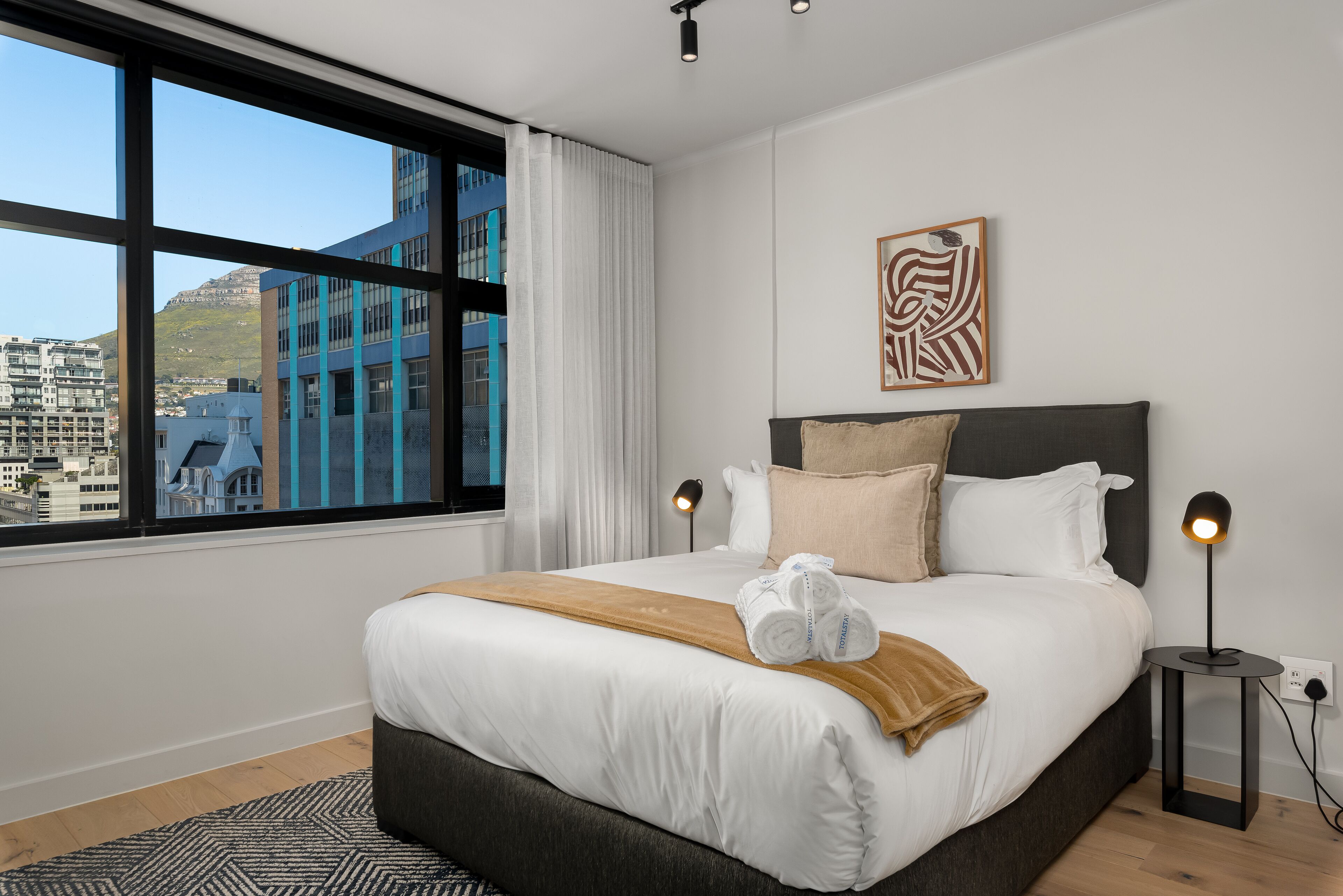 A modern hotel room with a large bed, a city view, and a unique wall art piece.