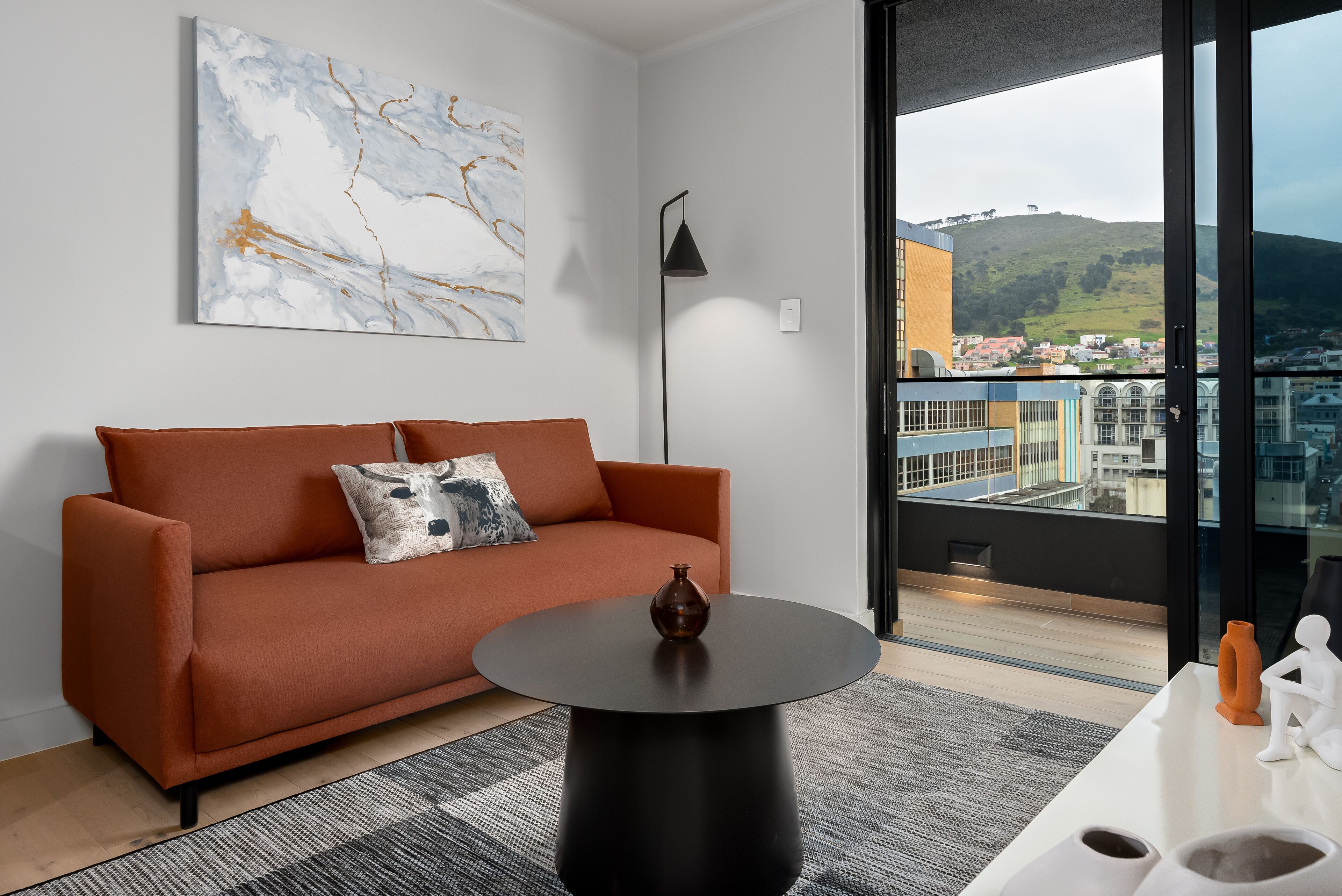 A modern living room with a sofa, a round black table, and a large window offering a city view.