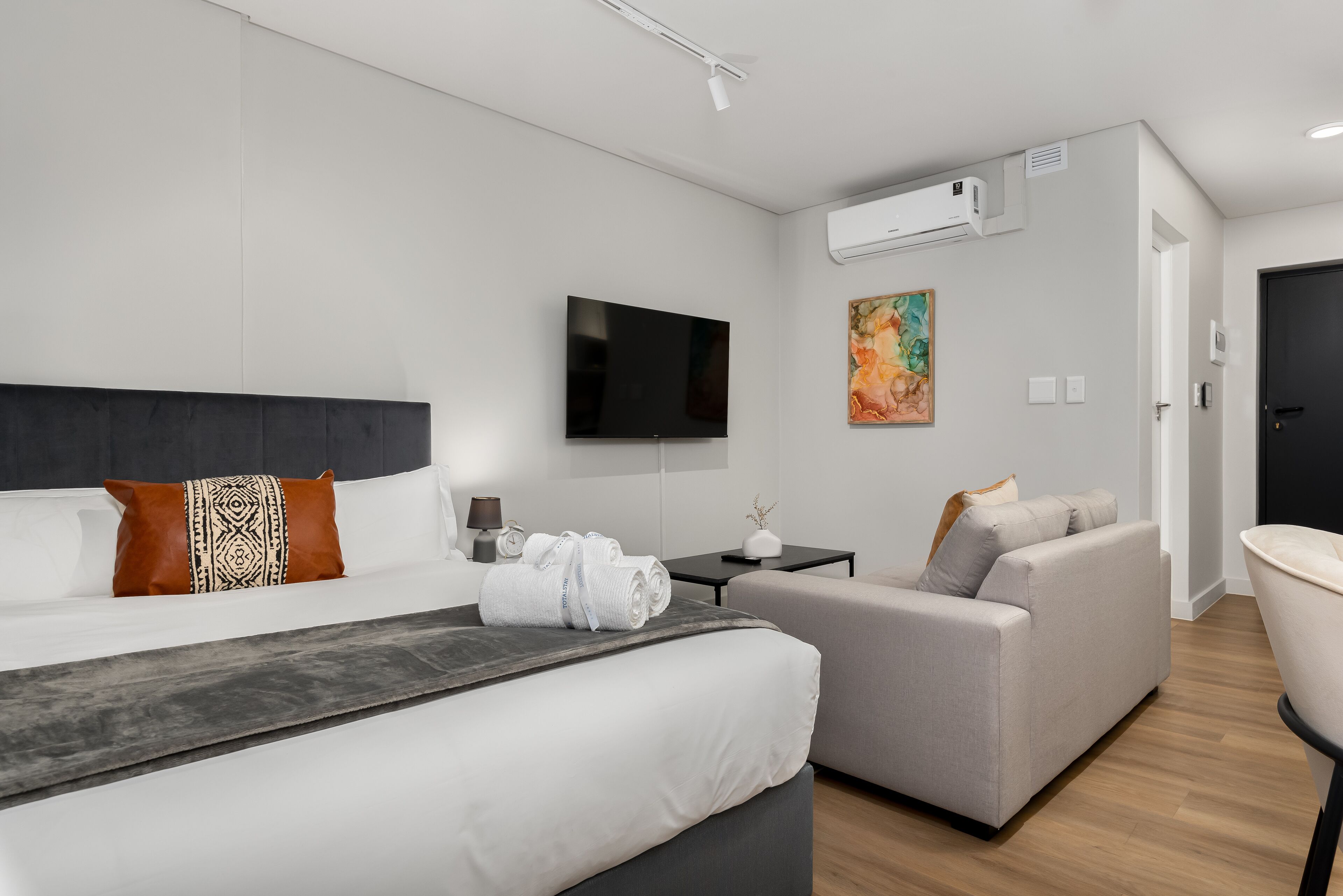 A modern hotel room with a large bed, a sofa, a flat-screen TV, and an air conditioning unit.