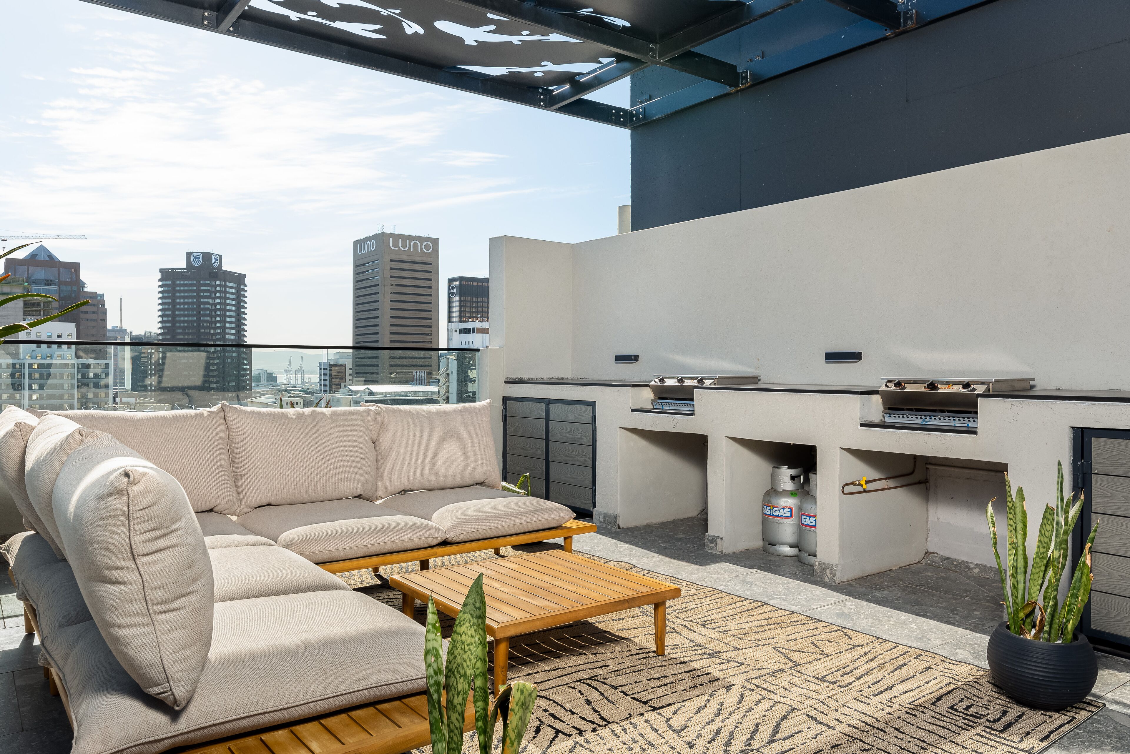 A rooftop terrace with a beige sectional sofa, a wooden coffee table, a grill, and a potted plant.