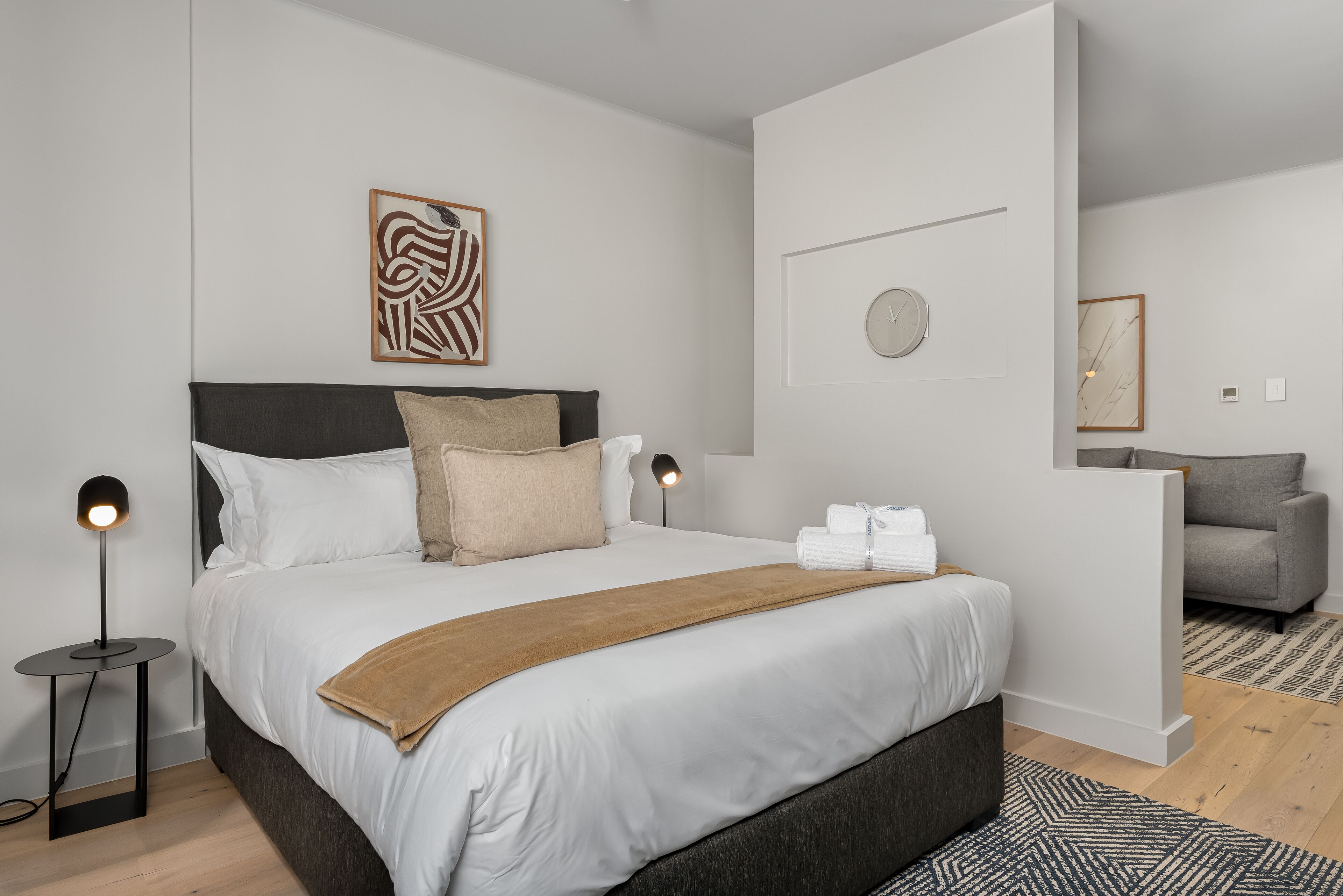 A modern bedroom with a large bed, a bedside lamp, a decorative wall piece, and a clock.