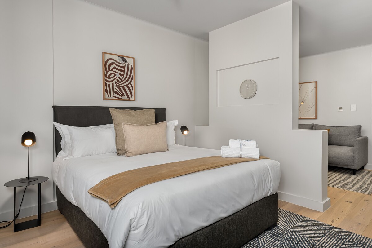 A modern bedroom with a large bed, a bedside lamp, a decorative wall piece, and a clock.
