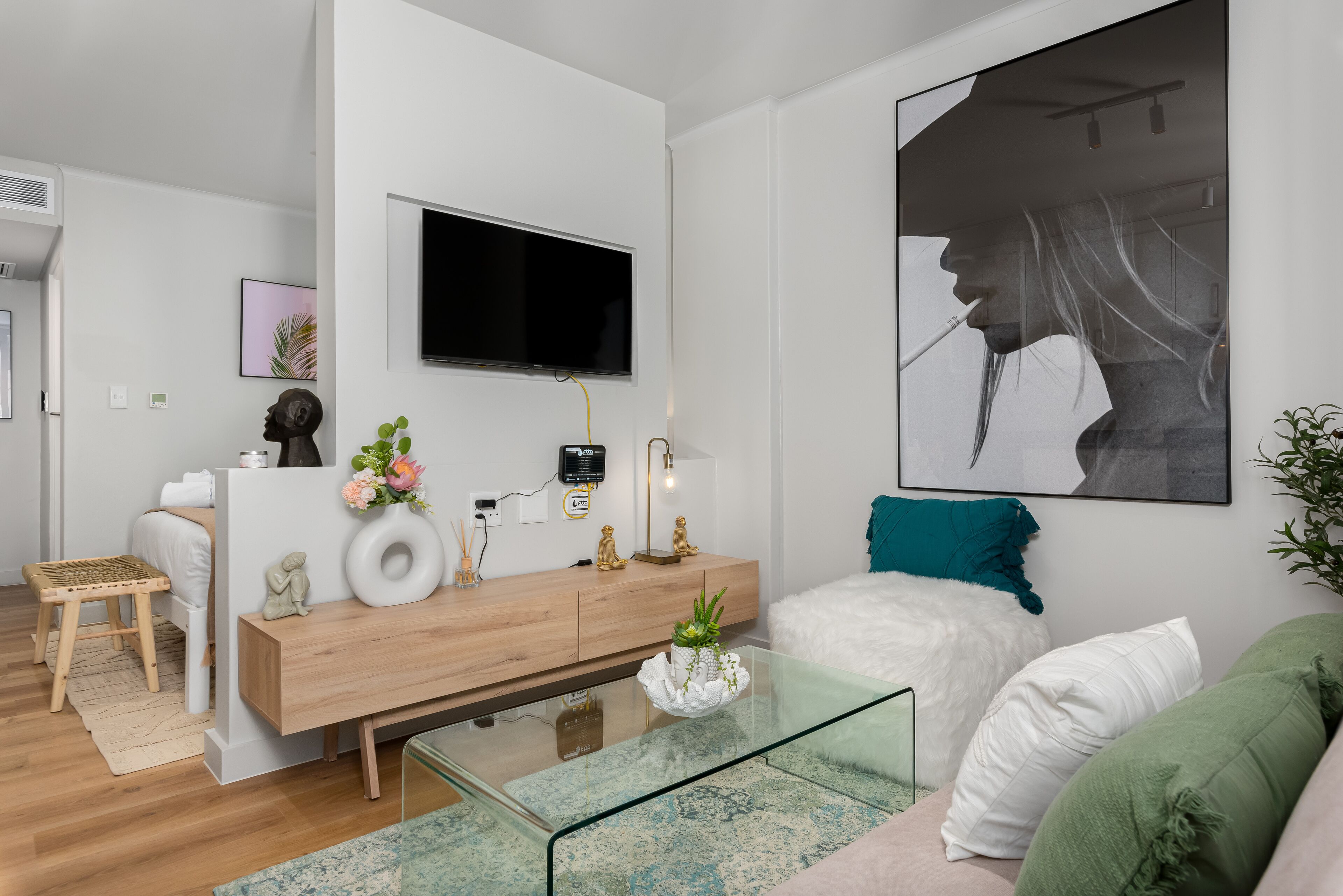 Una sala de estar moderna con una mesa de centro de vidrio, una consola de madera con objetos decorativos, un sofá blanco con cojines verde y blanco, y una obra de arte grande enmarcada en la pared.