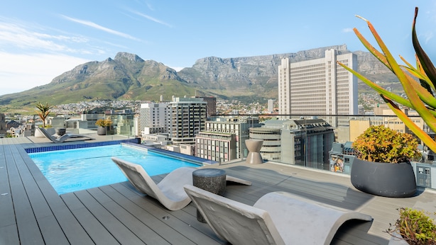 A rooftop pool with lounge chairs, a view of a cityscape, and mountains in the background.
