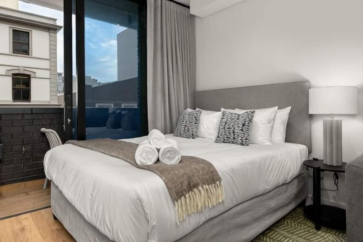 Een hotelkamer met een bed, kussens, een deken, een nachtkastje, een lamp en uitzicht.
