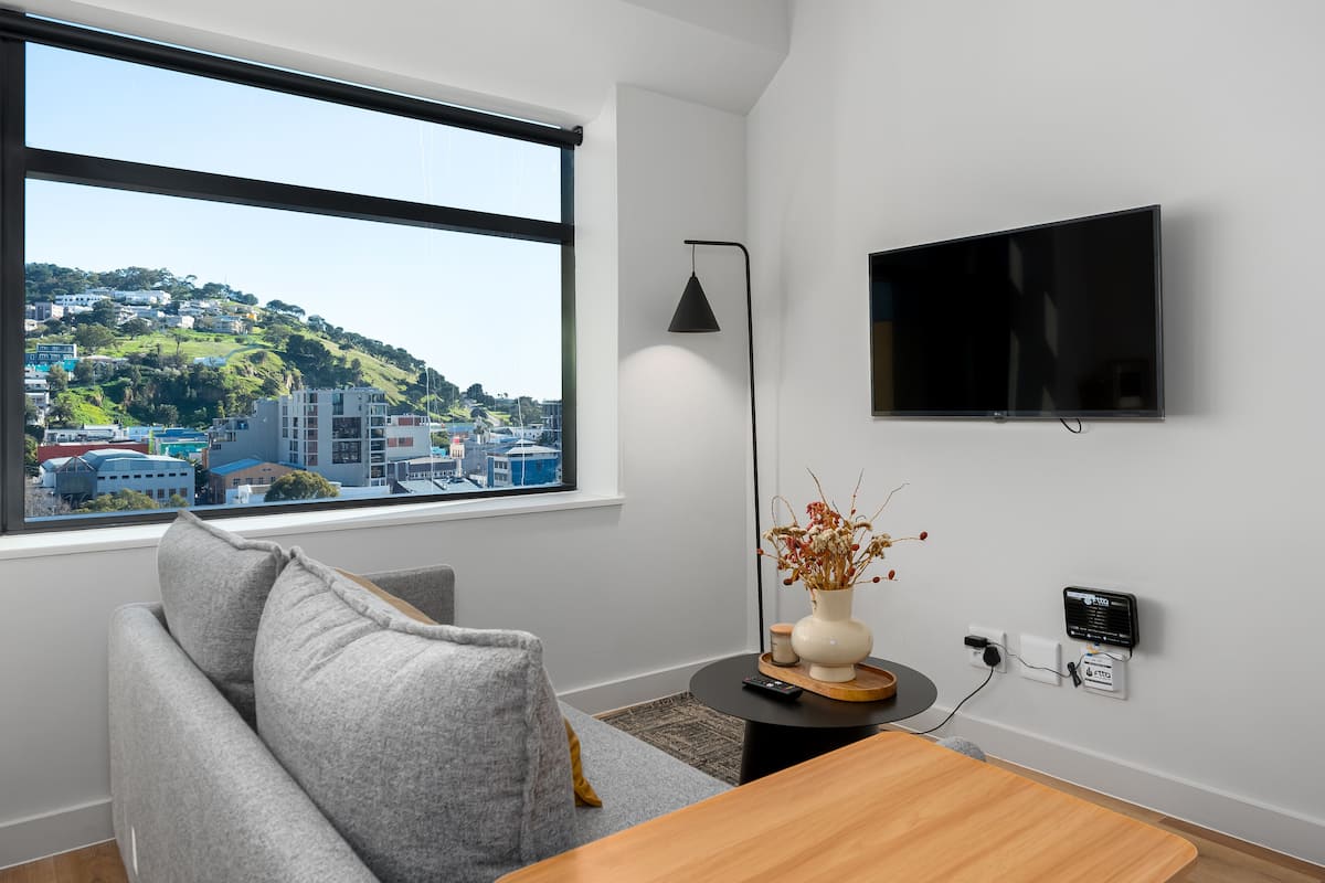 Een moderne woonkamer met een grijze bank, een houten salontafel, een vaas met gedroogde bloemen, een zwarte vloerlamp, een flatscreen televisie aan de muur en een raam met uitzicht op de stad.