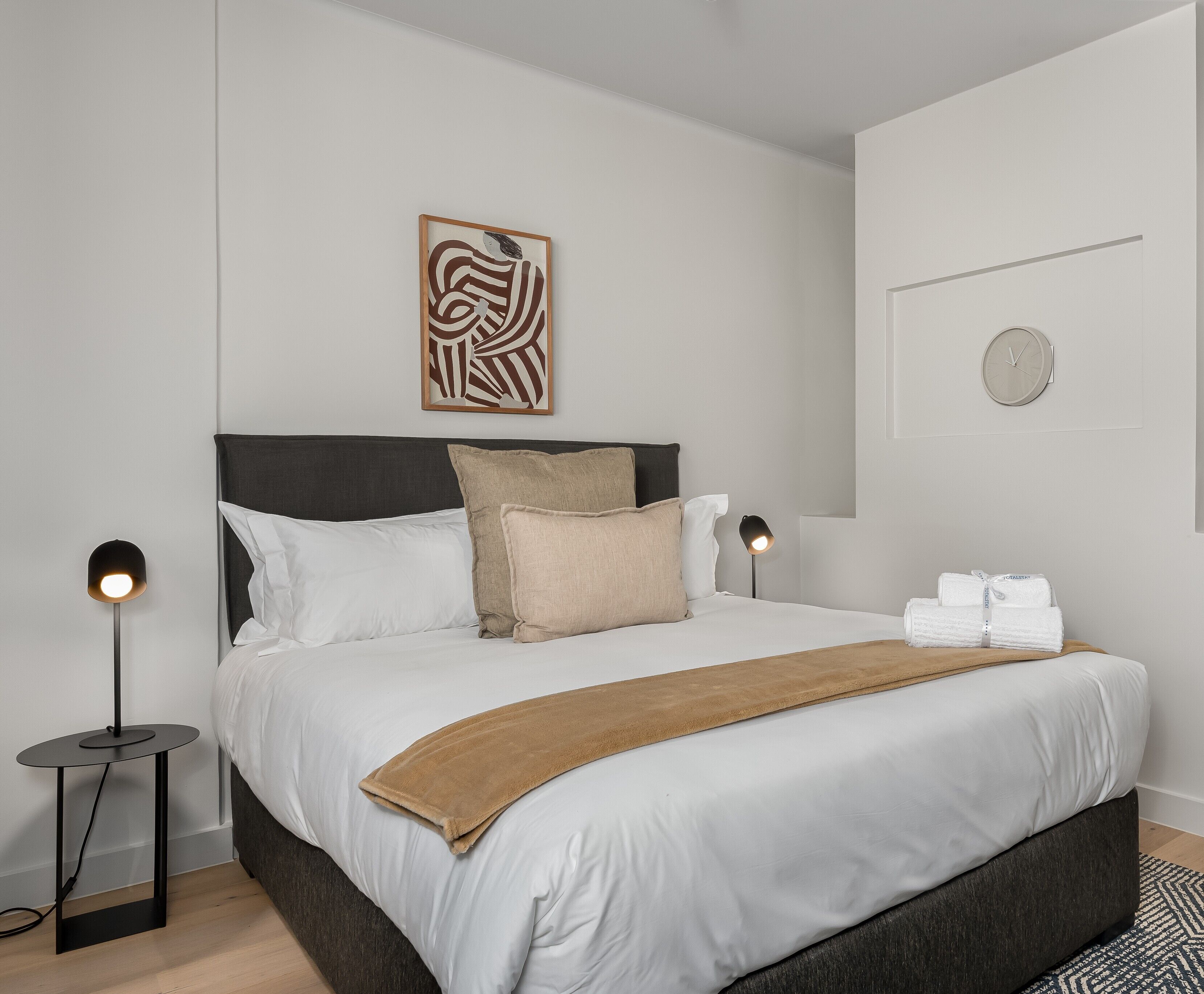 A modern hotel room with a large bed, a bedside table with a lamp, a wall clock, and a decorative piece above the bed.
