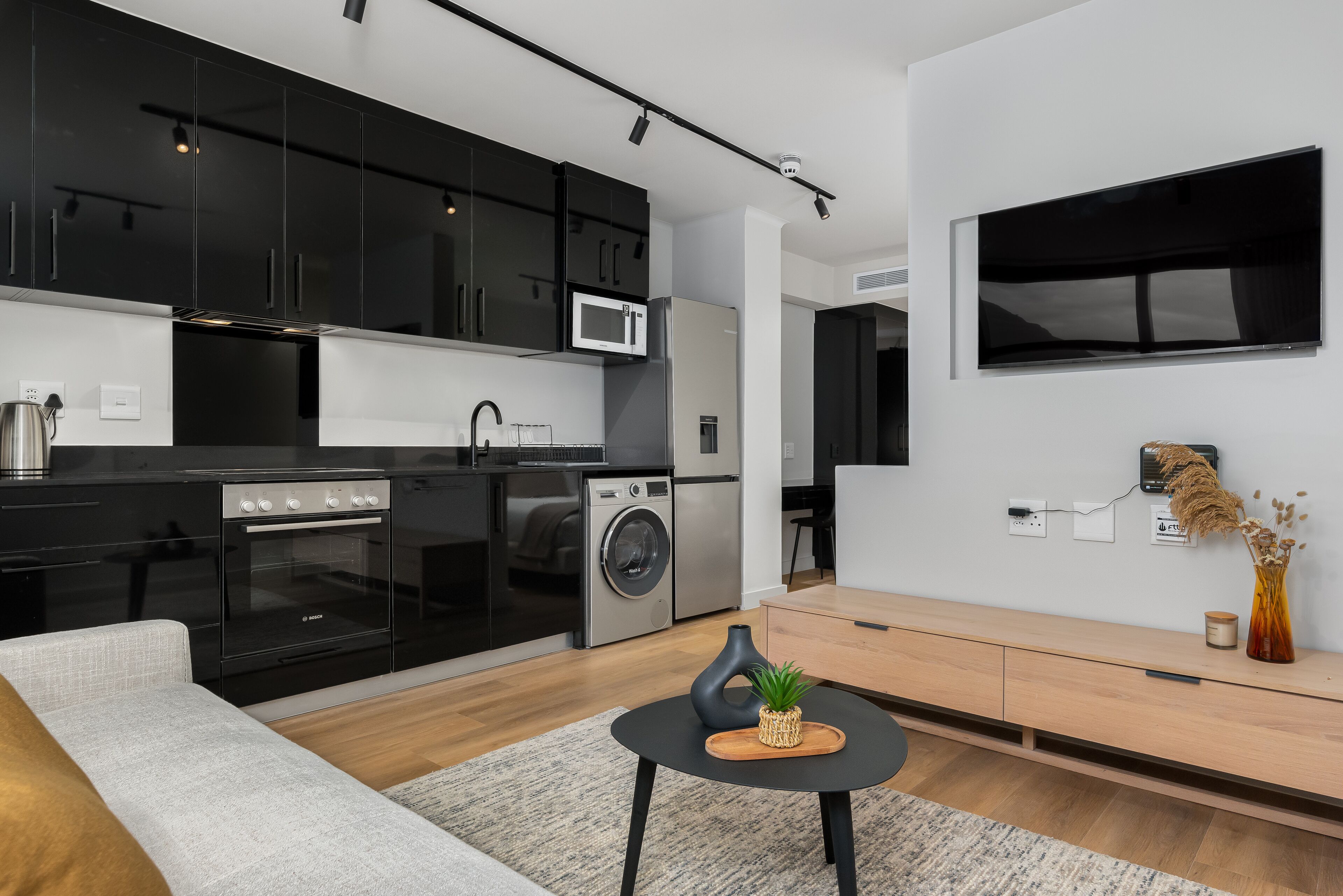 Una cocina moderna con armarios negros, un microondas y una lavadora. Hay una mesa de comedor de madera con una planta y una vela.