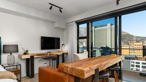 A modern hotel room with a large window, a wooden dining table, a sofa, a TV, and a small side table.