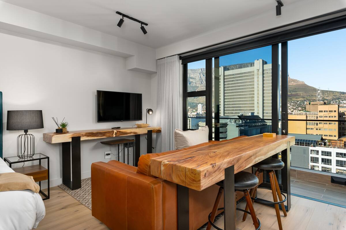 Een moderne hotelkamer met een groot raam, een houten eettafel, een bank, een televisie en een bijzettafeltje.