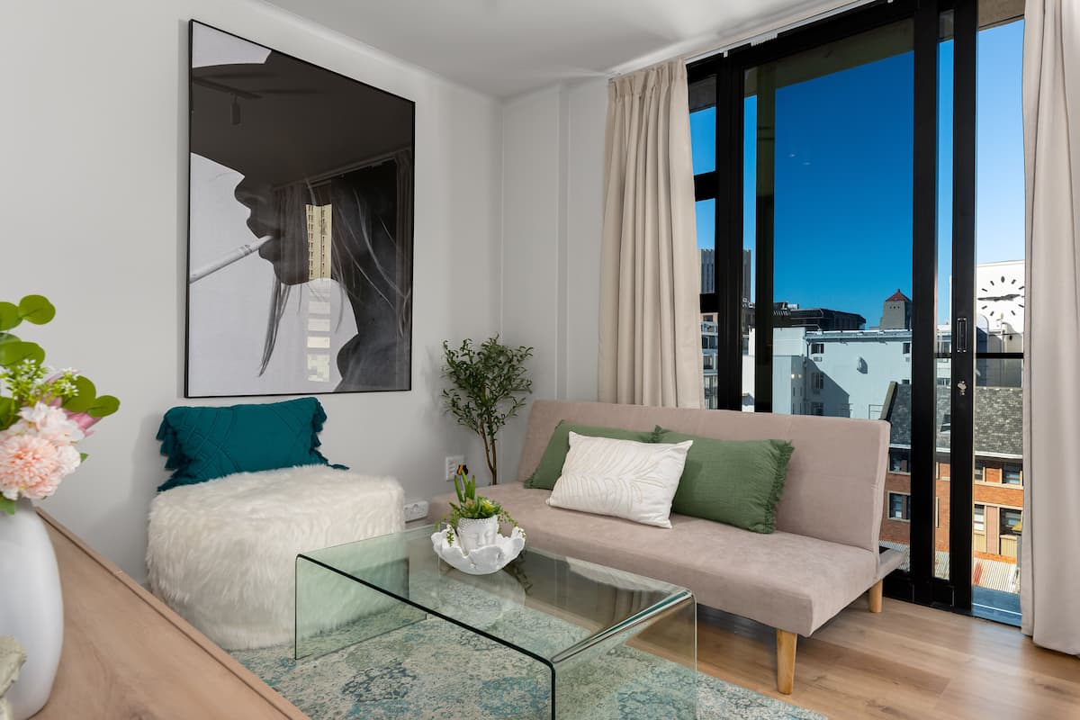 Een moderne woonkamer met een glazen salontafel, een bank met groene en witte kussens, een witte, pluizige fauteuil en een groot kunstwerk in een lijst aan de muur.