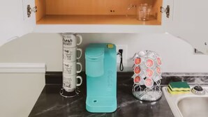 Ett kök med en bänkskiva, ett skåp med vinglas, en turkos vattenflaska, en kaffebryggare och en glasbehållare med svampar.