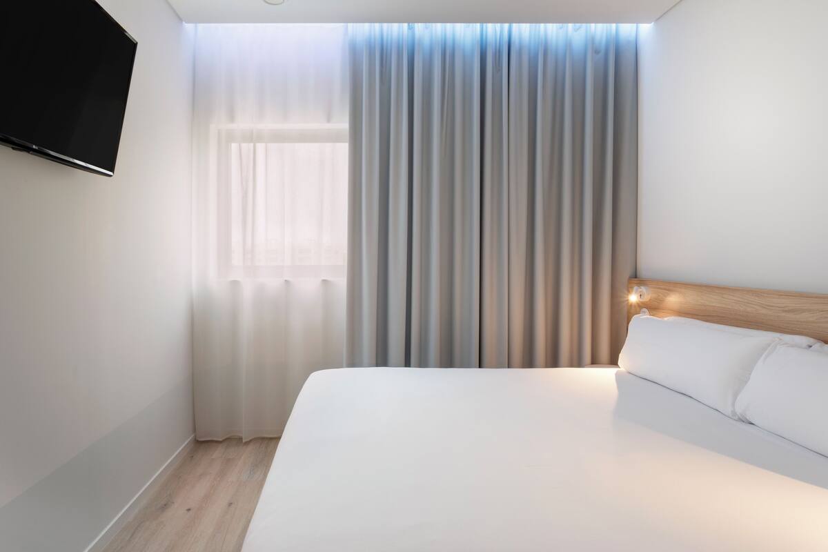 A hotel room with a large bed, a flat-screen TV mounted on the wall, and a window with sheer curtains.