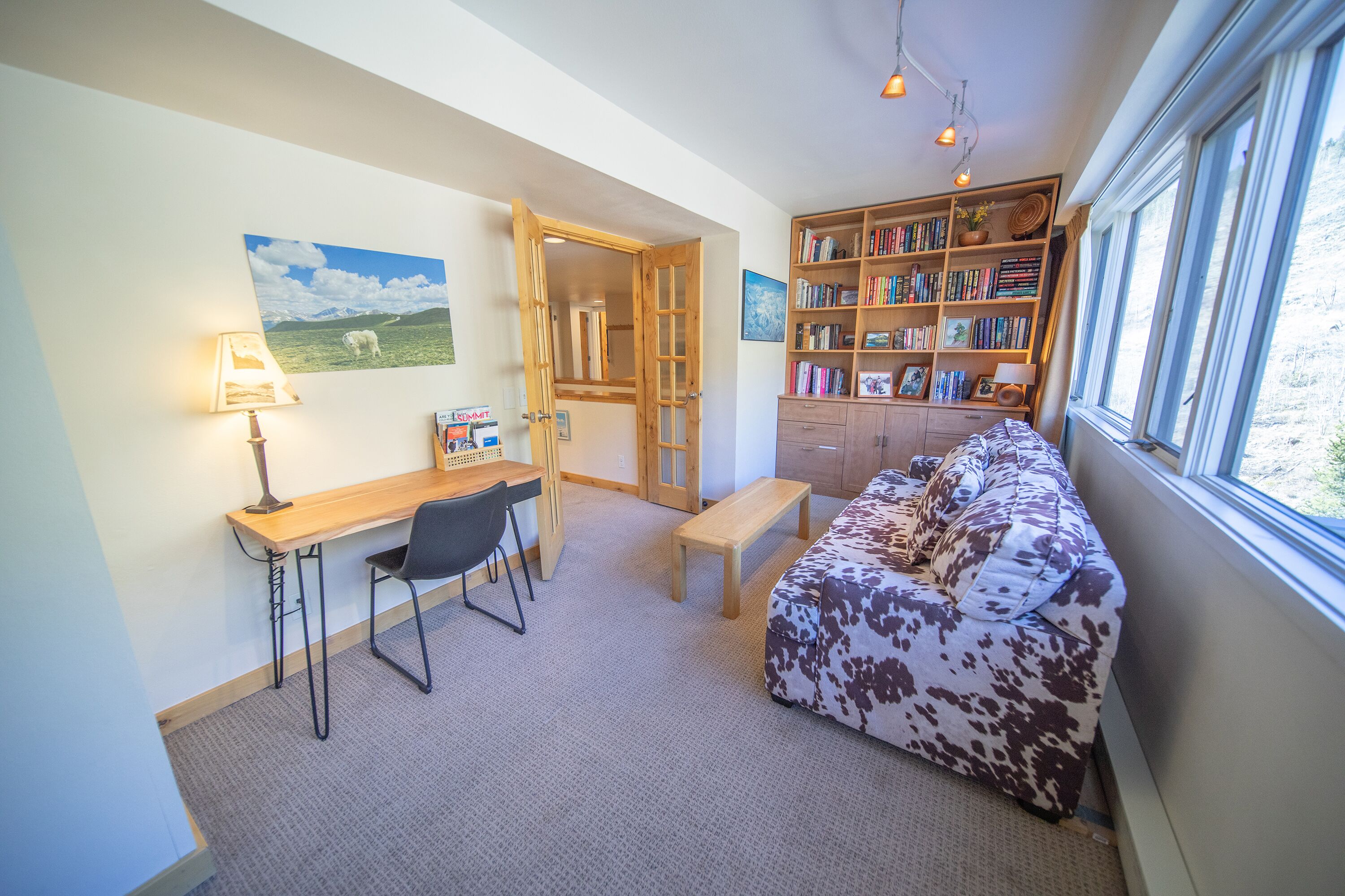 A cozy room with a wooden desk, a black chair, a cow-print sofa, a bookshelf, and a window with a view of the outdoors.