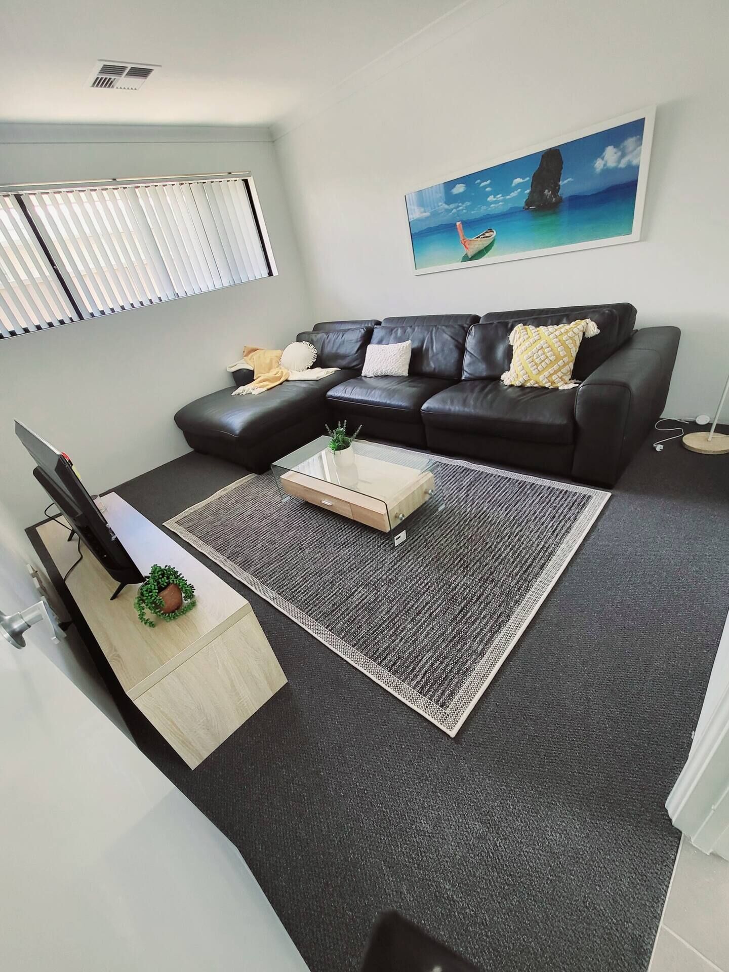 A modern living room with a black leather sofa, a glass coffee table, and a large painting of a coastal scene.