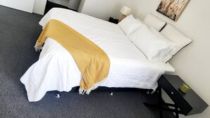 Een net opgemaakt bed met een witte dekbedovertrek en een gele plaid, een nachtkastje met een lamp, en een schilderij aan de muur.