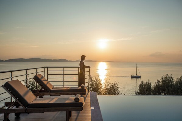 Une vue sur le coucher de soleil depuis une piscine à débordement, avec des chaises longues et une personne debout près de la balustrade.