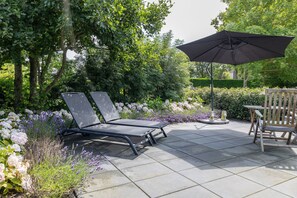Un patio al aire libre con dos sillones de descanso y una mesa, rodeado de abundante vegetación y plantas florecientes.