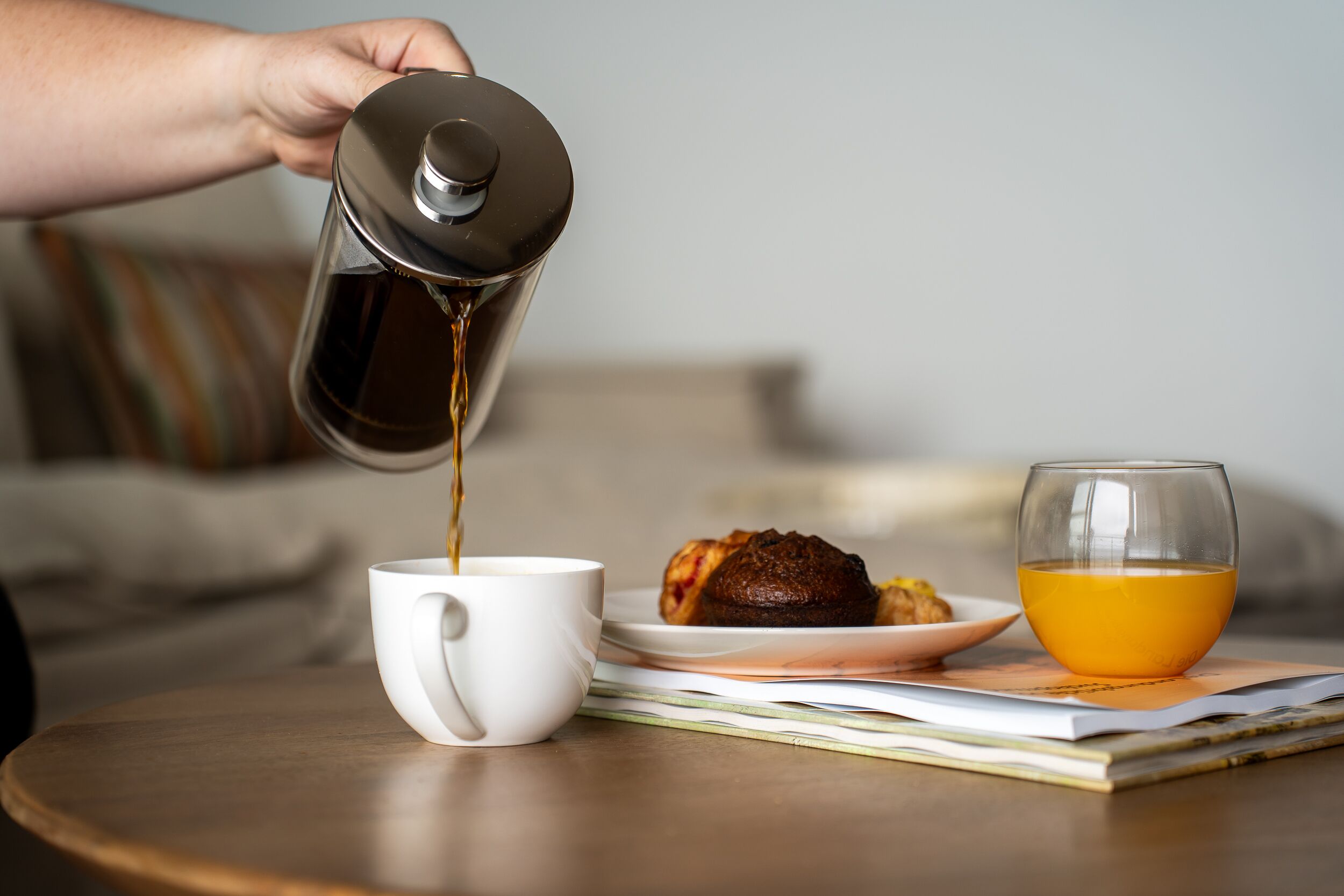 A hand pouring coffee into a white cup on a round wooden table, with a glass of orange juice and a plate with a pastry in the background.