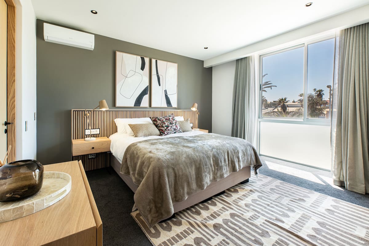 A modern hotel room with a large bed, a bedside table, a nightstand, and a view of the outdoors through a large window.