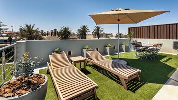 A rooftop terrace with wooden loungers, a table, and chairs under a large umbrella.