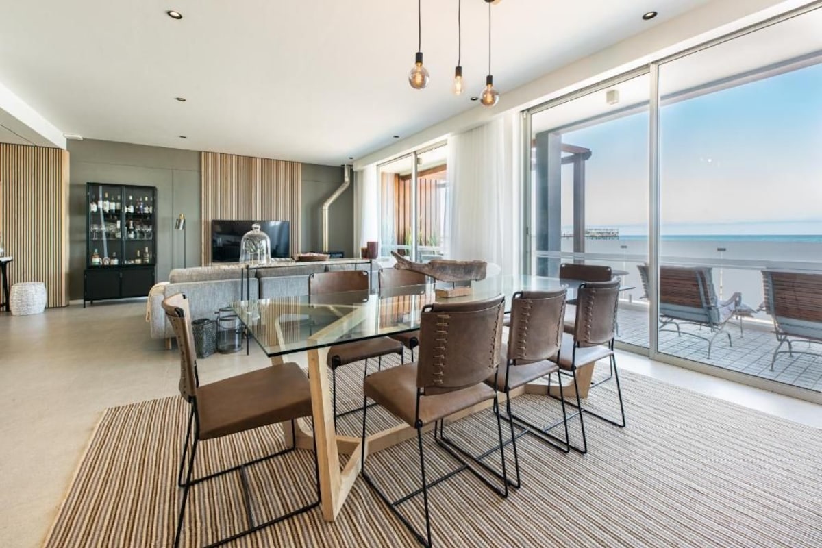 A modern kitchen with a glass dining table, chairs, and a view of the ocean.
