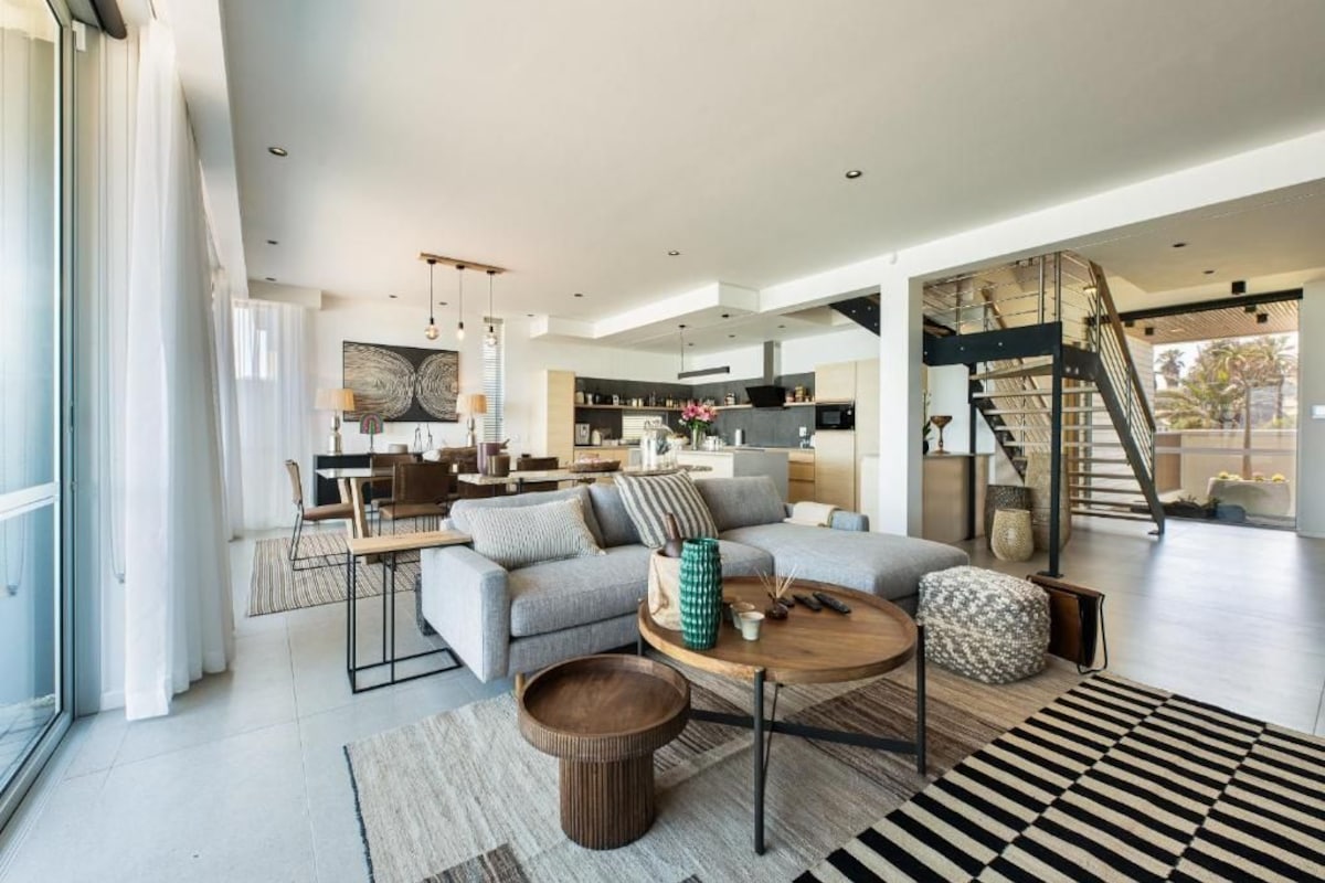 A spacious living area with a sofa, coffee table, and ottoman, a dining area with a table and chairs, and a kitchen in the background.