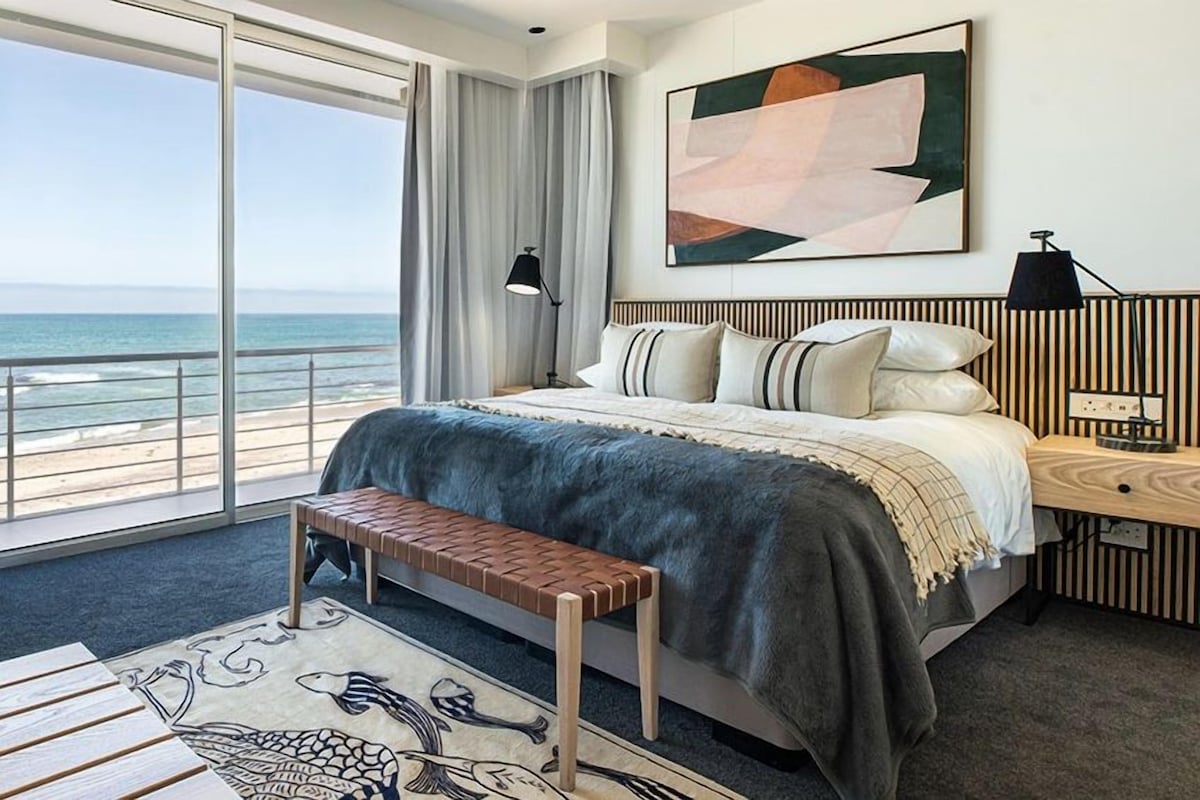 A bedroom with a large bed, a bench, a desk, and a view of the ocean.