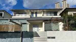 Une maison moderne à deux étages, dotée d’une façade en béton, d’un garage dont la porte est bien visible et d’un petit jardin agrémenté de galets blancs.