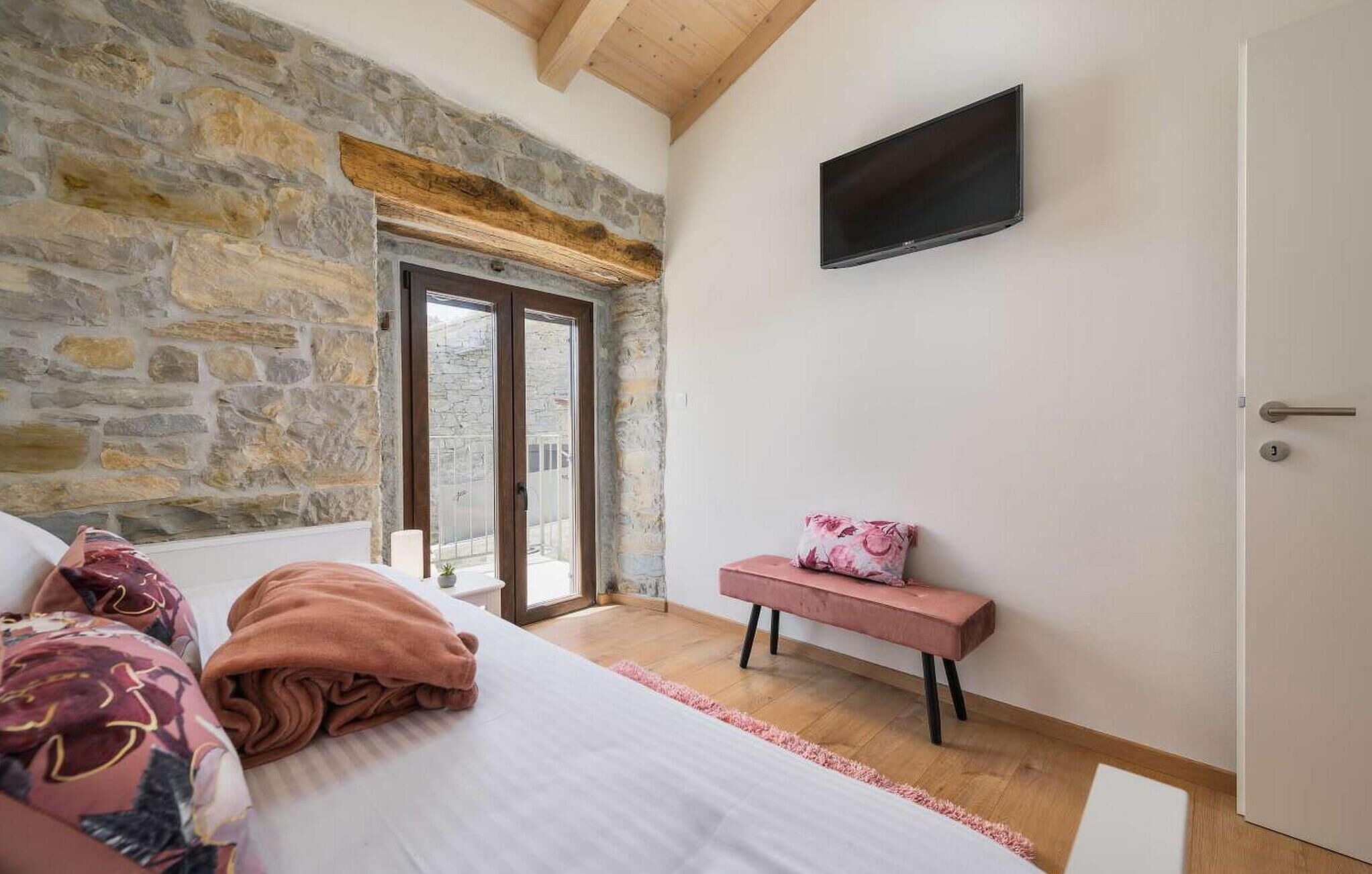 Un dormitorio con pared de piedra, cama con manta rosa, un banco rosa y televisor montado en la pared.