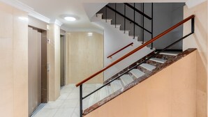 Un escalier moderne avec une rampe en bois et des balustrades en métal noir, menant à un ascenseur et à une pièce dont le mur est de couleur beige et le plafond, blanc.
