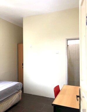 Une petite pièce, peu meublée, avec un lit simple, un bureau en bois et une chaise rouge.
