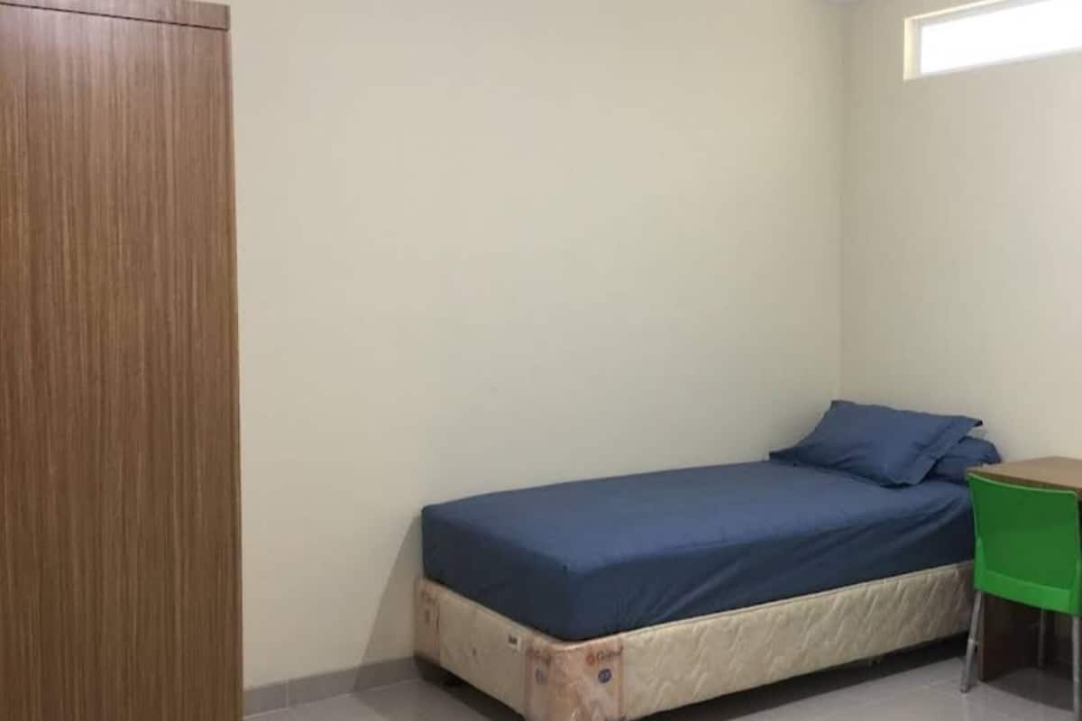 Une petite pièce, peu meublée, avec un lit simple, une armoire en bois et une chaise verte.