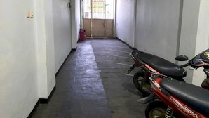 Un couloir étroit où est garée une moto, avec une poubelle rouge et une entrée fermée par un portail doté d’une fenêtre.