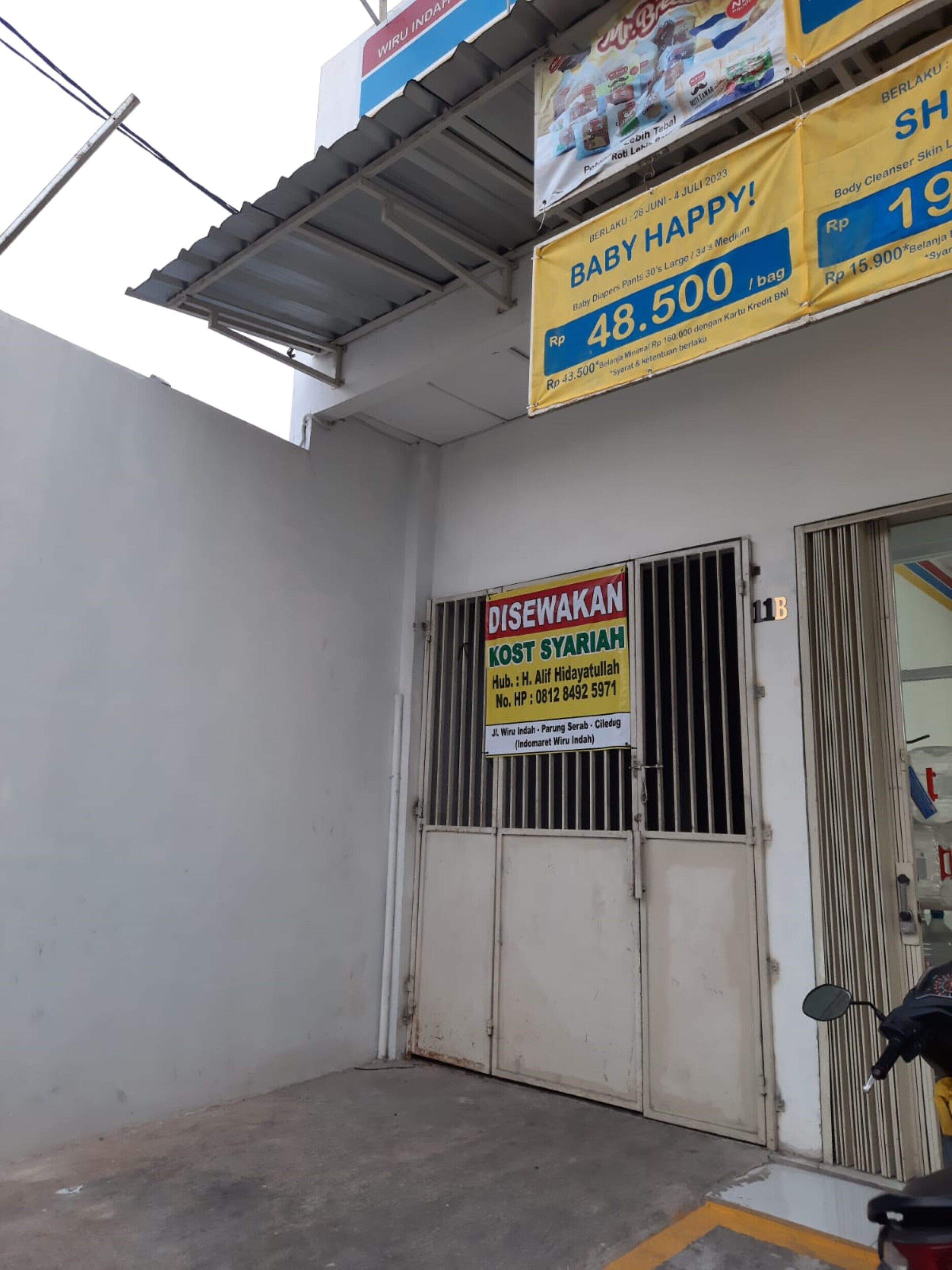 Fachada de una tienda que anuncia productos para bebés y una puerta con la etiqueta "DISEWAKAN KOST SYARIAH".