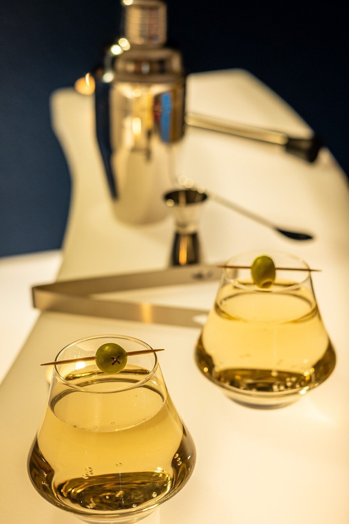 Two glasses of a yellowish liquid with olive garnishes on a white surface, a bar tool, and a bar bottle in the background.
