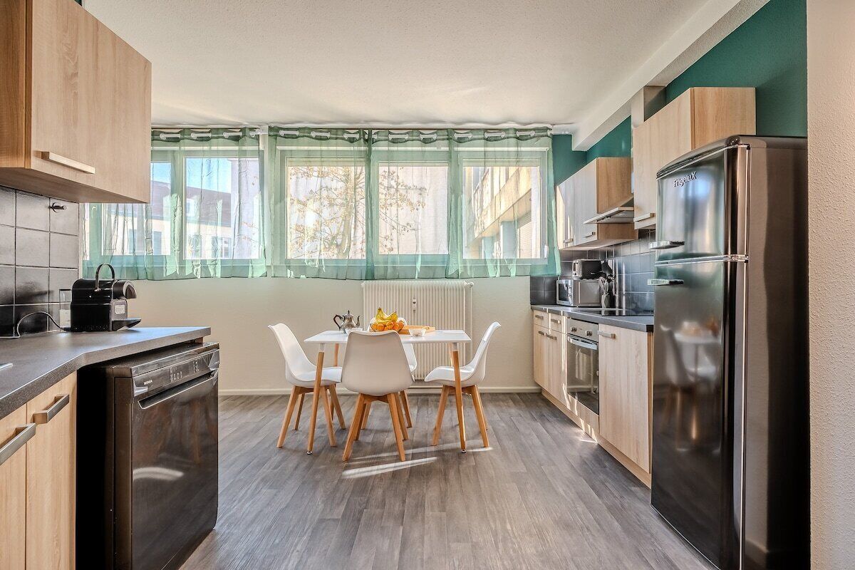 A compact kitchen with a dining table, chairs, a dishwasher, a refrigerator, and a window with curtains.