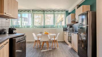 A compact kitchen with a dining table, chairs, a dishwasher, a refrigerator, and a window with curtains.