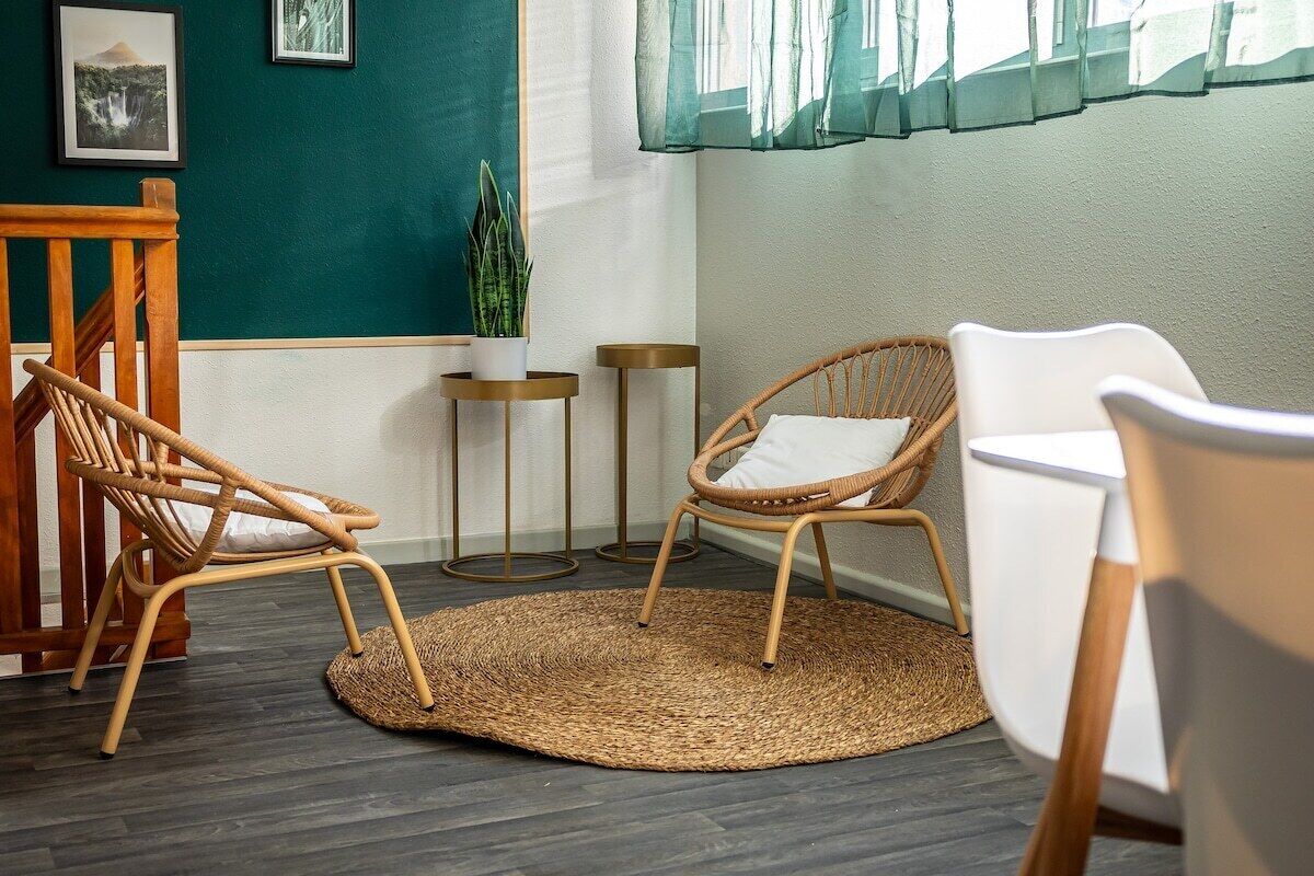 A room with a wooden chair, a white chair, a woven rug, and a small potted plant.