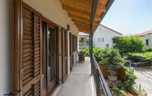 En balkon med træpersienner, et lille bord med stole og udsigt til en have og omkringliggende bygninger.
