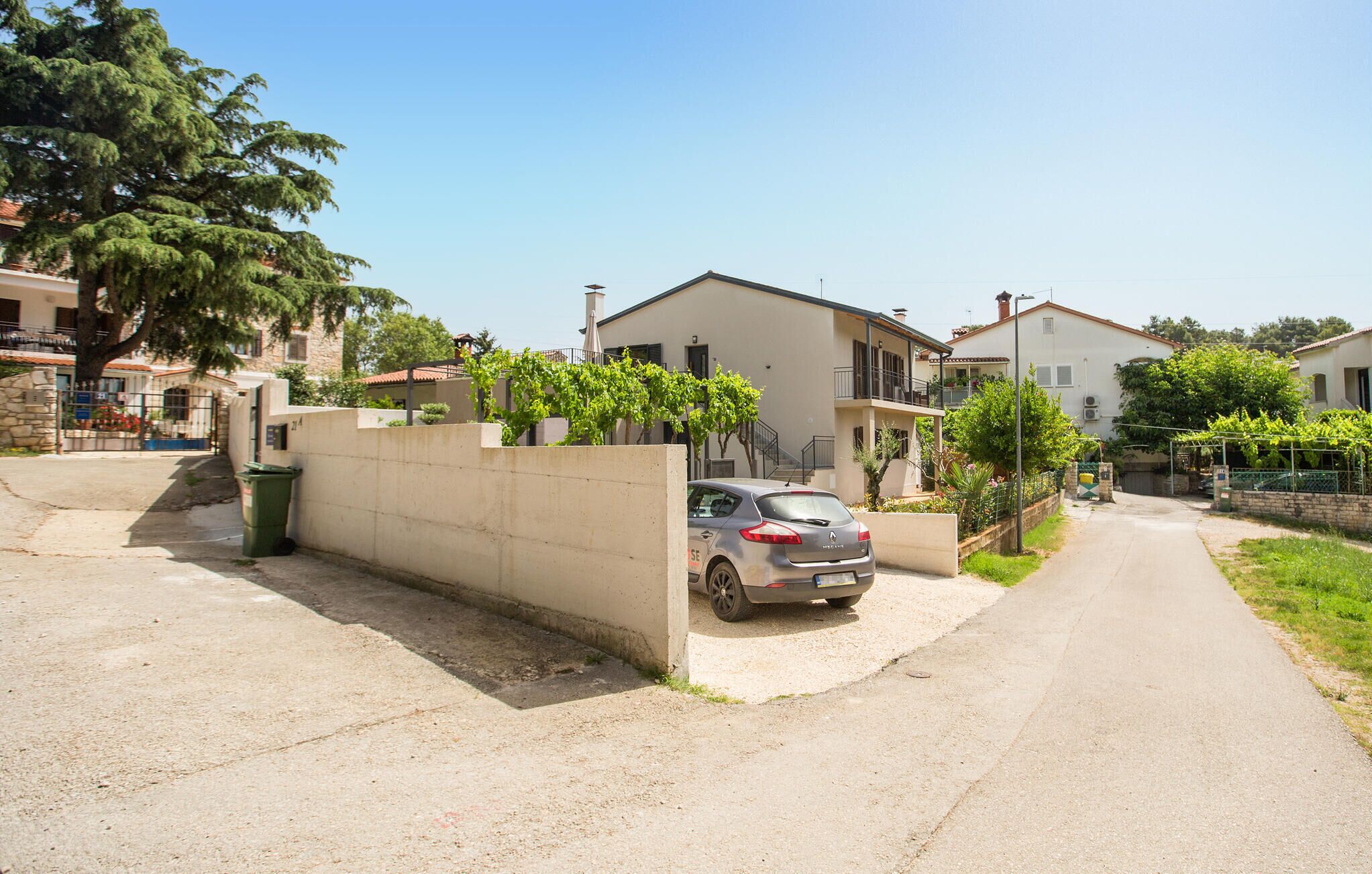 A residential street with houses, a parked car, and a concrete wall with greenery.