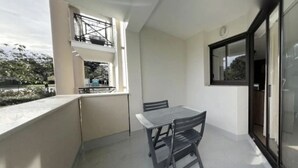 Un balcón con mesa y sillas, una ventana con vistas a árboles, y una puerta con panel de vidrio.