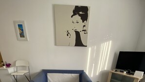 A room with a blue and white checkered bed, a white chair, a small table with a plant, a television on a stand, and a painting on the wall.